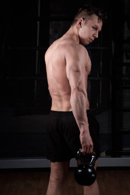 Çıplak gövdesi olan şişirilmiş adam spor salonunda duruyor, elinde bir kettlebell tutuyor ve omzunun üstündeki çerçeveye bakıyor.
