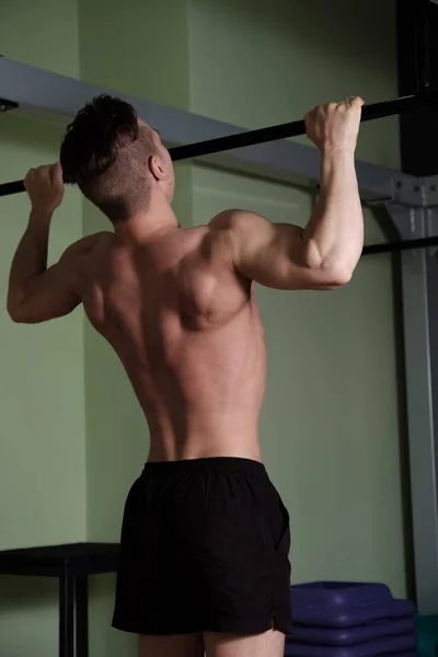 Un tipo musculoso con un torso desnudo se tira hacia arriba en la barra horizontal en el gimnasio, un tiro por la espalda . — Foto de Stock