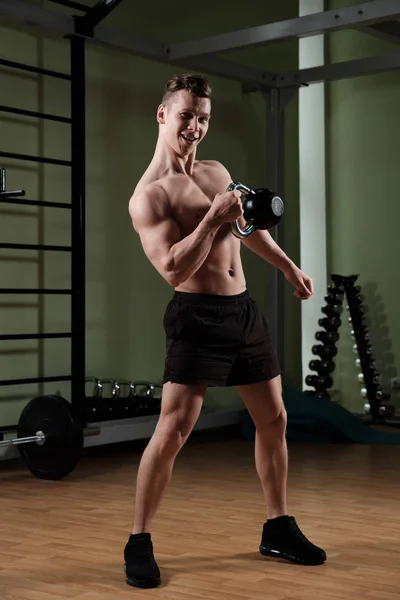Un tipo musculoso con un torso desnudo en pantalones cortos deportivos hace correr un kettlebell con una mano y mira a la cámara . —  Fotos de Stock