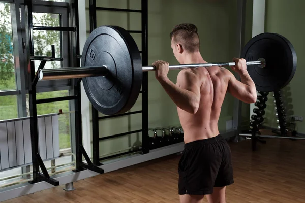 Muskulöser Typ mit nacktem Oberkörper in Sporthose steht mit einer Langhantel auf den Schultern in der Turnhalle und wird von hinten angeschossen. — Stockfoto