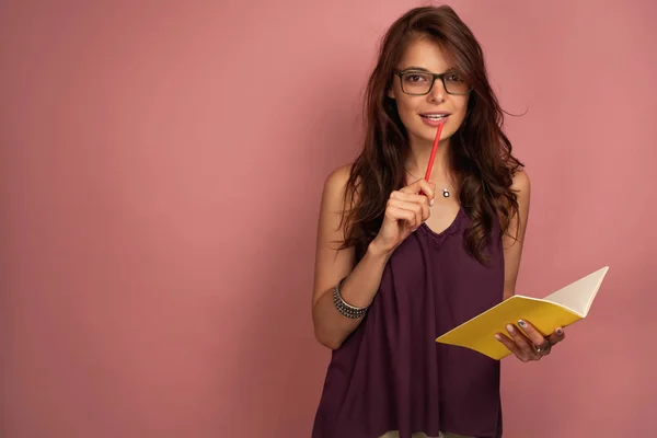 A sötét hajú lány egy lila tetején áll egy rózsaszín háttér egy sárga notebook és megnyomja a ceruzát a száját. — Stock Fotó