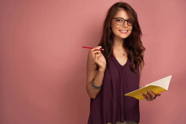 Mörkhårig flicka i en lila topp och glasögon med en anteckningsbok och penna står på en rosa bakgrund, ser glatt på kameran — Stockfoto