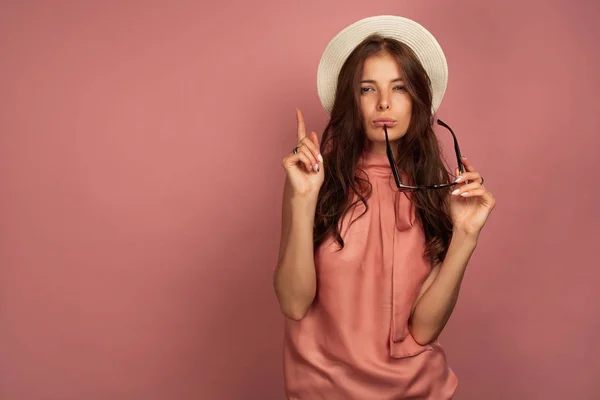 La chica de pelo oscuro con un vestido rosa, y un sombrero de paja sobre un fondo rosa, entrecerrando los ojos, señala su dedo a la parte superior . — Foto de Stock