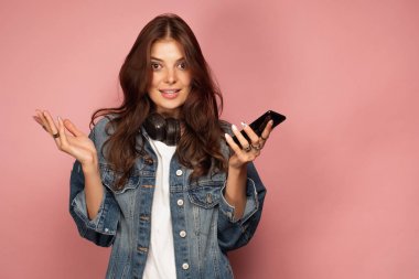 Kot pantolonlu bir kız boynunda kulaklık ile pembe bir arka plan üzerinde duruyor ve telefon ile el yayılır ve kameraya bakar.