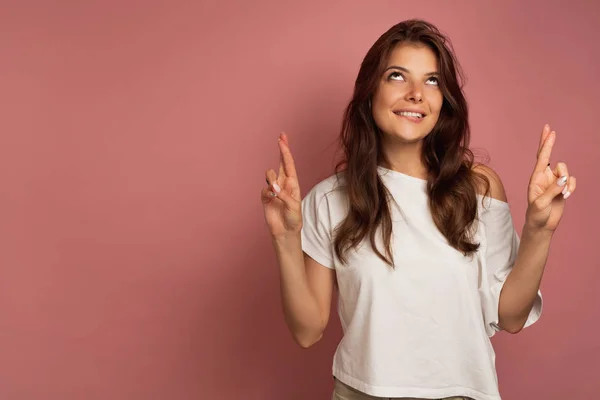 Ein dunkelhaariges Mädchen in weißem Top steht auf rosa Hintergrund, schaut nach oben, beißt in das Wrack und drückt die Daumen. — Stockfoto