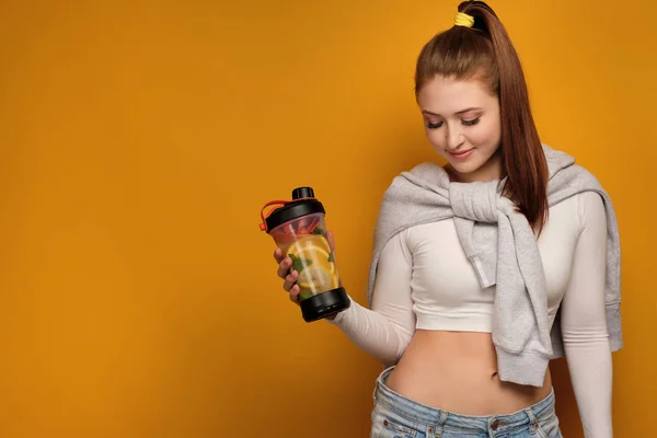 Het roodharige meisje staat op een gele achtergrond met haar hoofd naar beneden en glimlacht en houdt een sport Shaker in haar hand. — Stockfoto