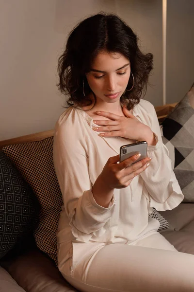 Fille bouclée s'assoit sur un canapé et regarde le téléphone avec sa main à sa poitrine . — Photo