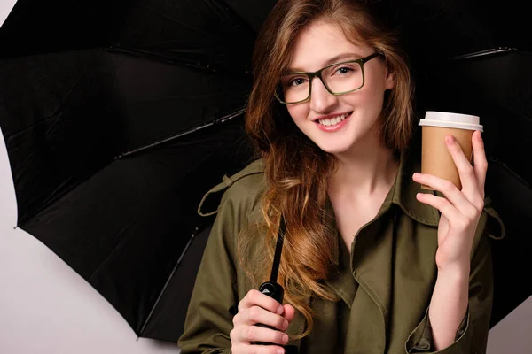 Una chica con un abrigo verde y gafas se levanta sobre un fondo blanco con un paraguas negro, sosteniendo una taza de papel en su mano . — Foto de Stock