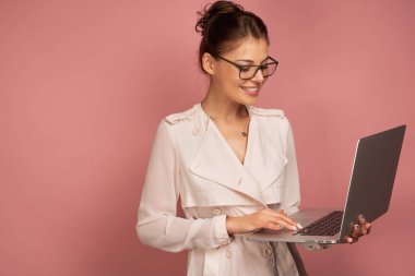 Açık ceket ve gözlük lü koyu saçlı bir kız bir dizüstü bilgisayar tutar ve gülümseyerek monitör bakar.