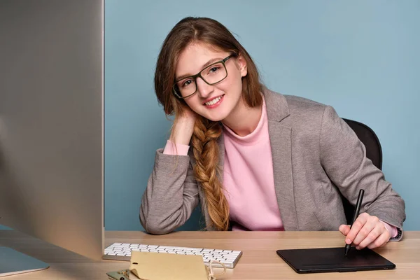 Egy lány egy rózsaszín pulóvert, szürke kabát és szemüveg ül egy számítógép és mosolyog a kamera. — Stock Fotó