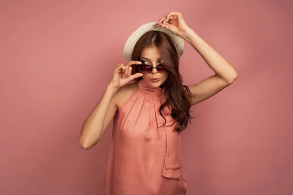 A sötét hajú lány egy rózsaszín ruhában áll egy rózsaszín háttér, emelő kalapját, és kihagyom napszemüveg, nézett le az oldalára. — Stock Fotó