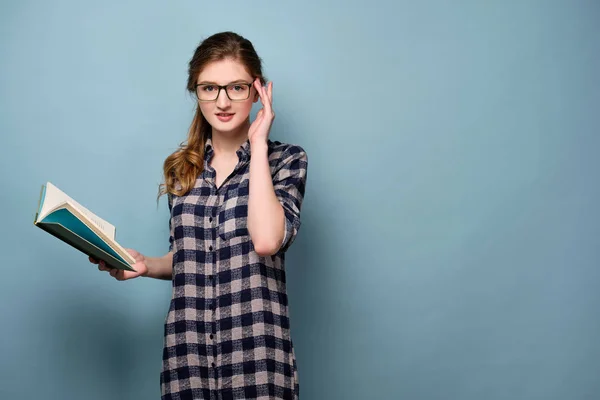 En ung flicka i en pläd klänning och glasögon står på en blå bakgrund med en öppen bok i handen och tittar på kameran. — Stockfoto