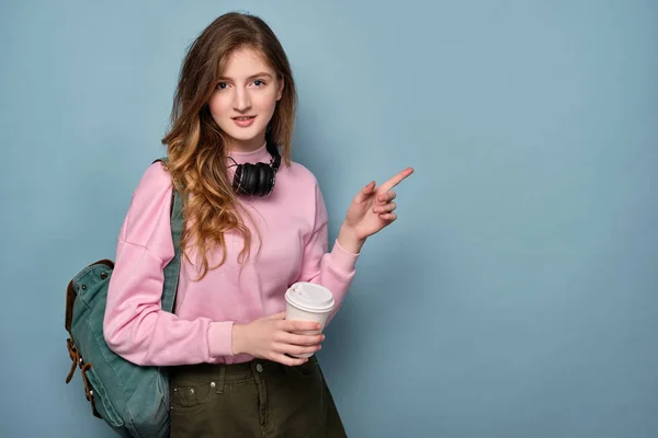 분홍색 스웨터와 배낭을 든 헤드폰을 입은 소녀가 손에 플라스틱 컵을 들고 서서 손가락을 옆으로 가리킵니다.. — 스톡 사진