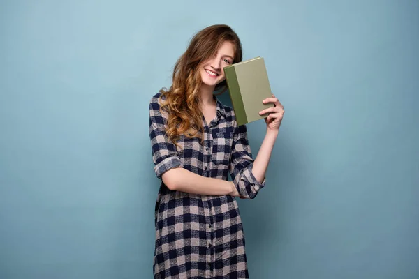 Egy fiatal lány egy kockás inget áll a kék háttér mögé bújva egy könyvet. — Stock Fotó