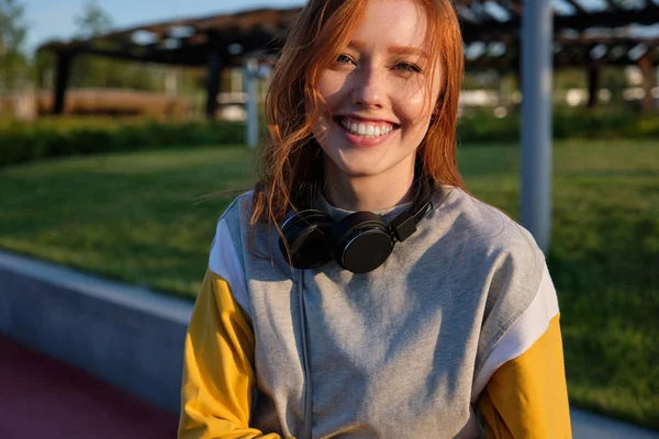 Redhead flicka med fräknar och gröna ögon med hörlurar på halsen står på gatan och ler brett i ramen — Stockfoto