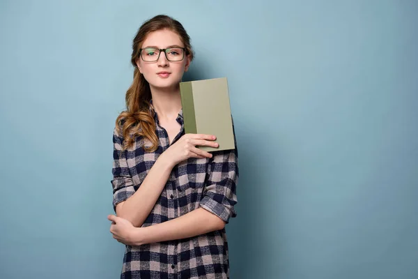 En ung flicka i en pläd skjorta och glasögon står på en blå bakgrund med en grönbok i handen. — Stockfoto