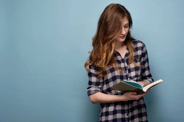 En ung flicka i en pläd skjorta står på en blå bakgrund och ser ner i en bok med huvudet böjt. — Stockfoto
