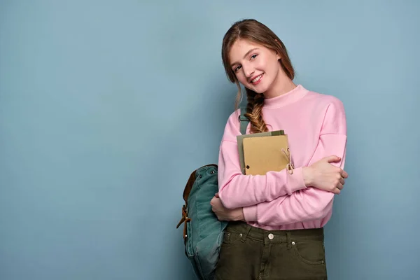 En flicka i en rosa tröja med ryggsäck och står i en omfamning med böcker och leende på kameran. — Stockfoto