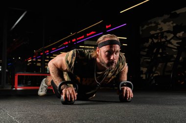Spor üniformalı kaslı adam, elleriyle kettlebell'e yaslanmış bir barda duruyor.