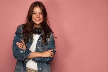 Kot pantolon lacivert kız boynunda kulaklık ile pembe bir arka plan üzerinde duruyor ve kollarını göğsüne çapraz gülümsüyor