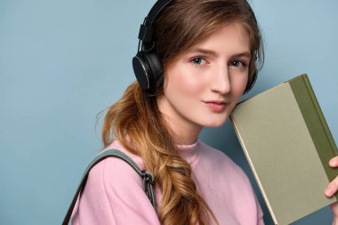 Pembe kazak ve sırt çantası ile kulaklık lı genç bir kız mavi bir arka plan üzerinde duruyor ve yüzünün önünde bir kitap tutar.
