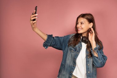 Kot pantolonlu koyu saçlı bir kız boynunda kulaklıklarla pembe bir arka plan üzerinde duruyor ve gülümserken selfie çekiyor.