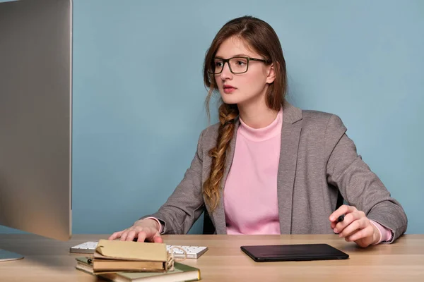 En flicka i en rosa tröja, en grå jacka och glasögon sitter vid ett bord, noga tittar på bildskärmen. — Stockfoto