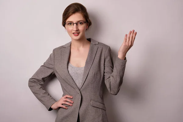 Una chica con una chaqueta gris y gafas se coloca sobre un fondo blanco, mira a la cámara, levantando la mano hasta la parte superior . — Foto de Stock