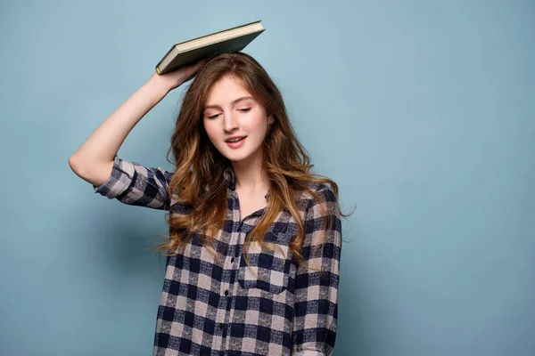 En ung flicka i en pläd skjorta står på en blå bakgrund och ser ner med en bok på huvudet. — Stockfoto