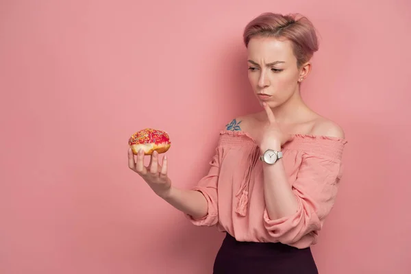 En flicka med kort rosa hår, håller en munk i handen, och rynkar pannan på honom, på en rosa bakgrund. — Stockfoto