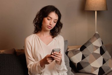Koyu saçlı kıvırcık bir kız yastıkların arasında oturur ve elindeki telefona bakar..