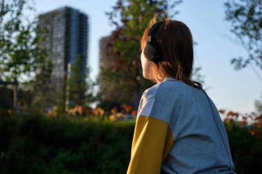 Kulaklıklı kızıl saçlı kız şehrin arka planında gün batımı nda sokakta oturuyor, arkadan çerçeve