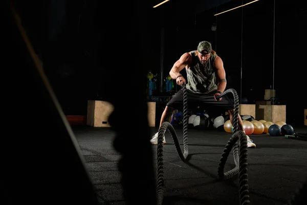 Muscular man in a sports uniform and a baseball cap performs an exercise with ropes in the gym — Stock Photo, Image