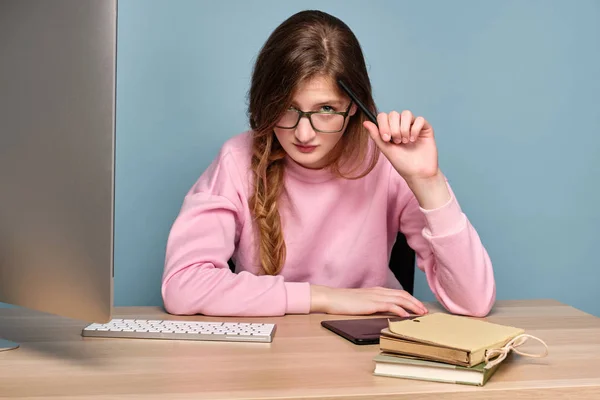 분홍색 스웨터를 입은 소녀가 컴퓨터와 함께 테이블에 앉아 펜으로 머리를 긁고 카메라의 안경을 바라보고 있습니다.. — 스톡 사진