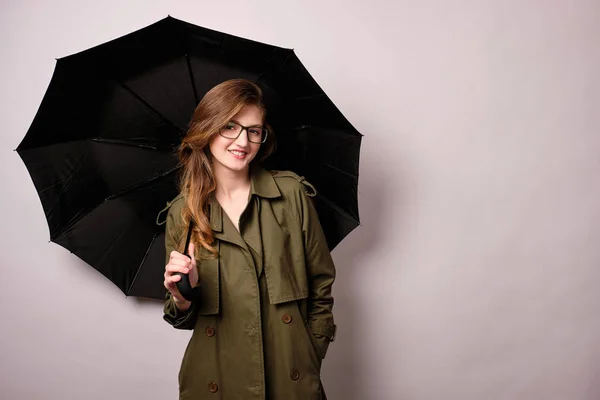 Uma menina loira de óculos e capa de chuva está de pé com um guarda-chuva preto e sorrindo para a câmera com a mão no bolso — Fotografia de Stock
