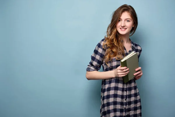 En ung flicka i en pläd klänning står på en blå bakgrund och håller en bok i händerna och ler mot kameran. — Stockfoto