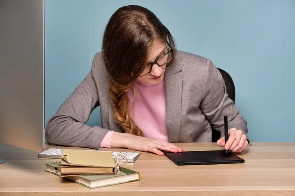 분홍색 스웨터, 회색 재킷과 안경을 쓴 소녀가 테이블에 앉아 머리를 숙이고 전자 태블릿에 그립니다.. — 스톡 사진