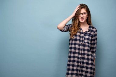 Ekose elbise ve gözlüklü genç bir kız mavi bir arka plan üzerinde duruyor ve ağzı açık sürpriz kamera görünüyor.