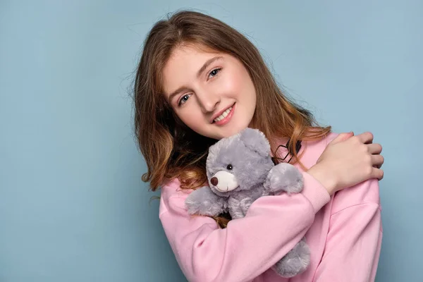 A girl in a pink sweater stands ona blue background and smiling, presses a bear toy to her.