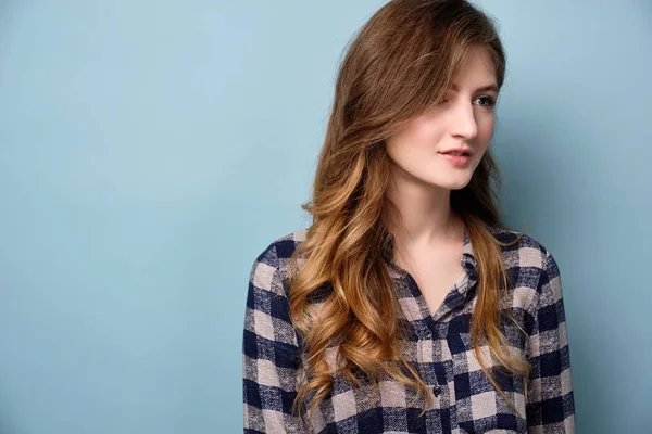 Uma jovem com uma camisa xadrez fica em um fundo azul e olha para o lado . — Fotografia de Stock