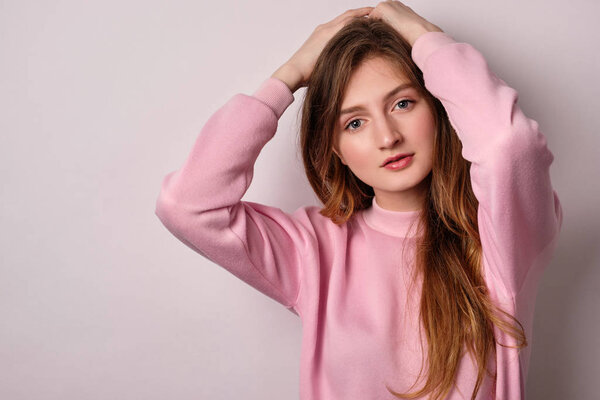 Блондинка в розовой толстовке стоит на белом фоне и смотрит в рамку с руками на голове
