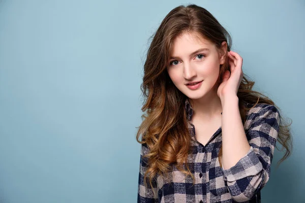 Een jong meisje in een geruite overhemd staat tegen een blauwe achtergrond en kijkt naar de camera, borstelen haar haar achter haar oor. — Stockfoto