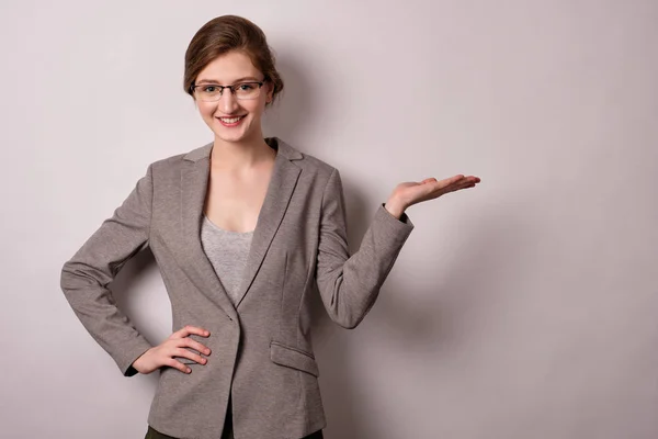 Chica rubia en gafas y una chaqueta gris con soportes sobre un fondo blanco, apunta con la palma a un lado y sonríe — Foto de Stock