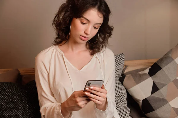 Açık bluzlu koyu saçlı kıvırcık bir kız yastıkların arasına oturur ve elindeki telefona bakar..