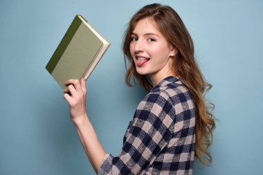 Ekose elbiseli bir kız mavi bir arka plan üzerinde kameraya bakarak duruyor ve elinde bir kitap tutarak, dilini dışarı sopa.