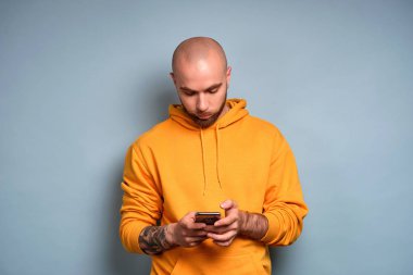 Sarı tişörtlü sakallı bir adam mavi bir arka plan üzerinde duruyor, başını eğiyor, elindeki telefona bakıyor.