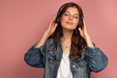 Kot pantolonlu sevimli bir kız, gözleri kapalı müzik izlerken zevkle pembe bir arka plan üzerinde duruyor.