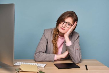 Pembe kazaklı, gri ceketli ve gözlüklü bir kız masaya oturmuş, üzgün bir şekilde kameraya bakıyor, elleriyle başını destekliyor..