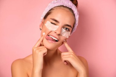 Saçları toplanmış leylaklı bir kız gözlerinin altında bantlarla dikiliyor ve dilini çıkarıp göz kırpıyor..