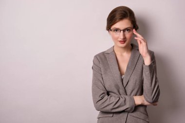 Gri ceketli bir kız beyaz arka planda duruyor, kameraya bakıyor ve gözlüklerini düzeltiyor..